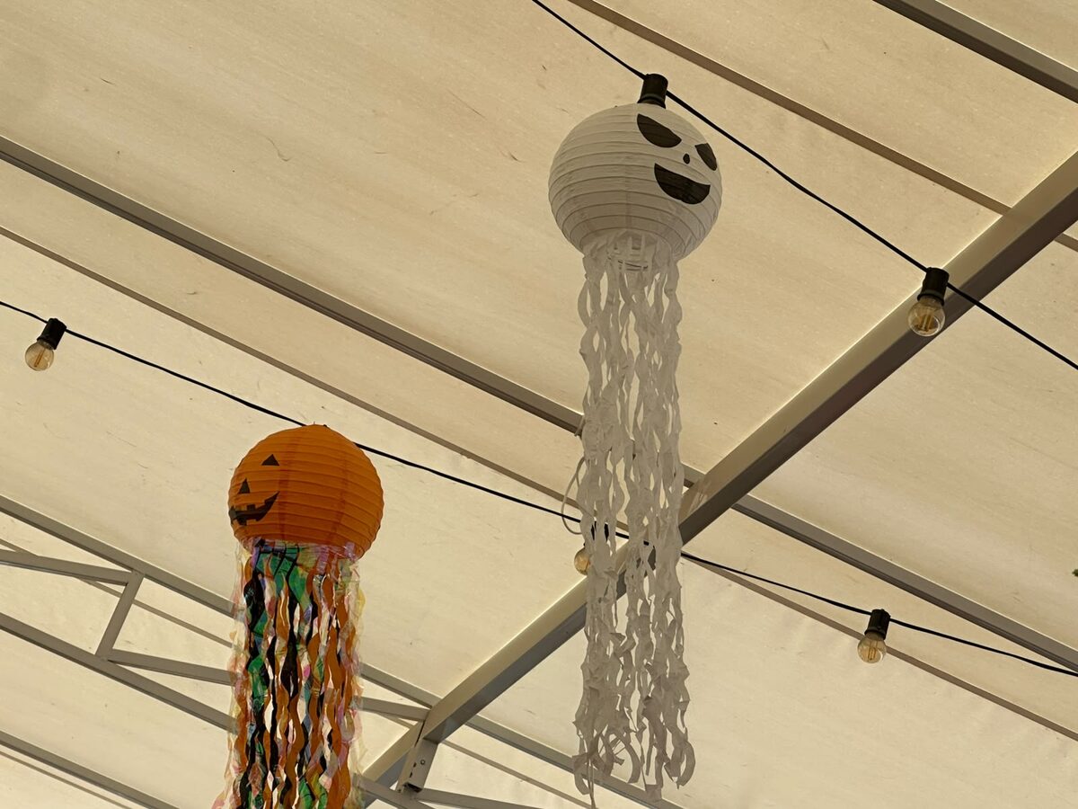 Lámparas colgantes de Halloween con caras de fantasma y calabaza, hechas de papel con tiras de flecos, sobre el techo de la terraza de Las Puertas con guirnaldas de bombillas cálidas