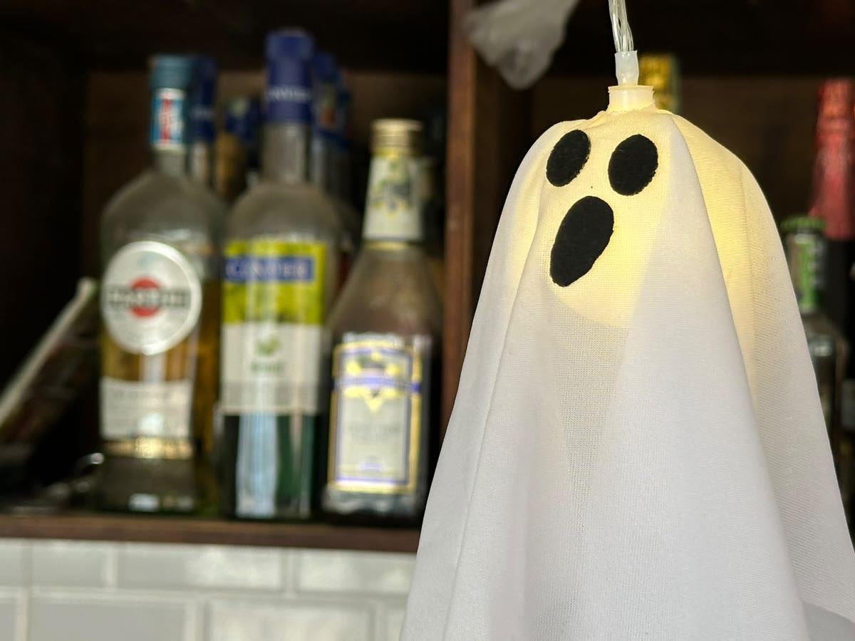 Fantasma de tela iluminado desde adentro colgando sobre la barra de madera de Las Puertas, con botellas de licor desenfocadas al fondo