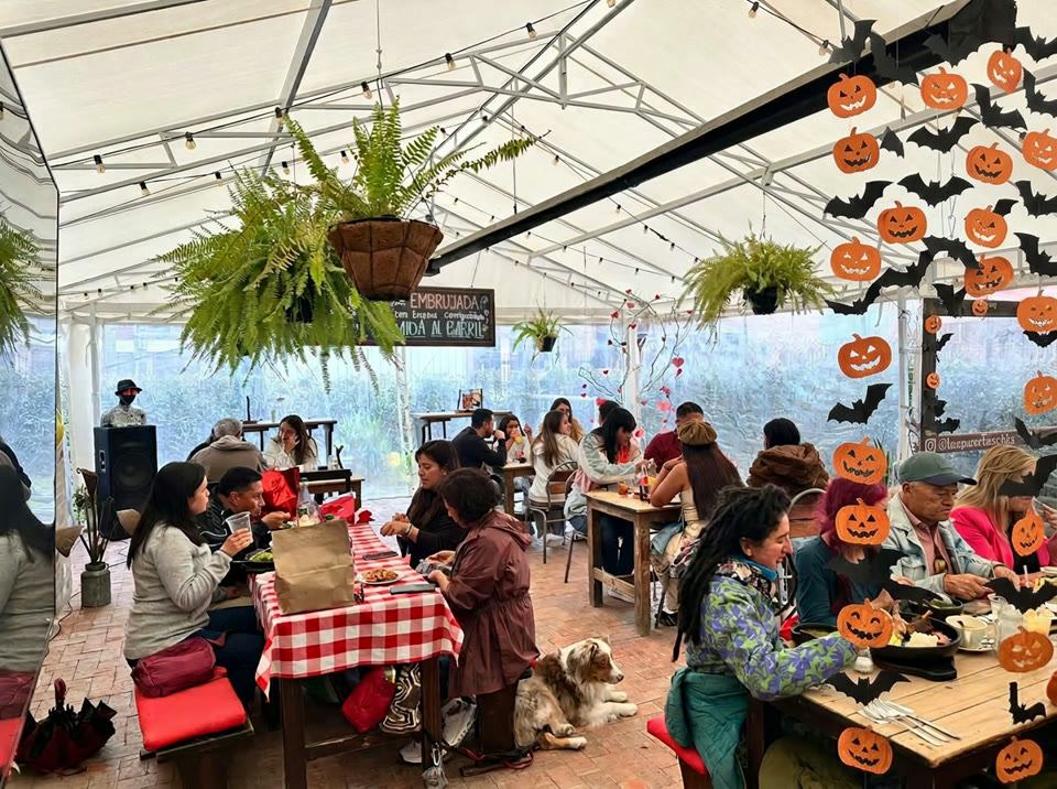 Terraza de Las Puertas en Chía: mesas rústicas de madera bajo techo de lona con guirnaldas de luces, comensales compartiendo platos al barril, un pizarrón que dice EMBRUJADA y un marco decorado con calabazas y murciélagos de Halloween