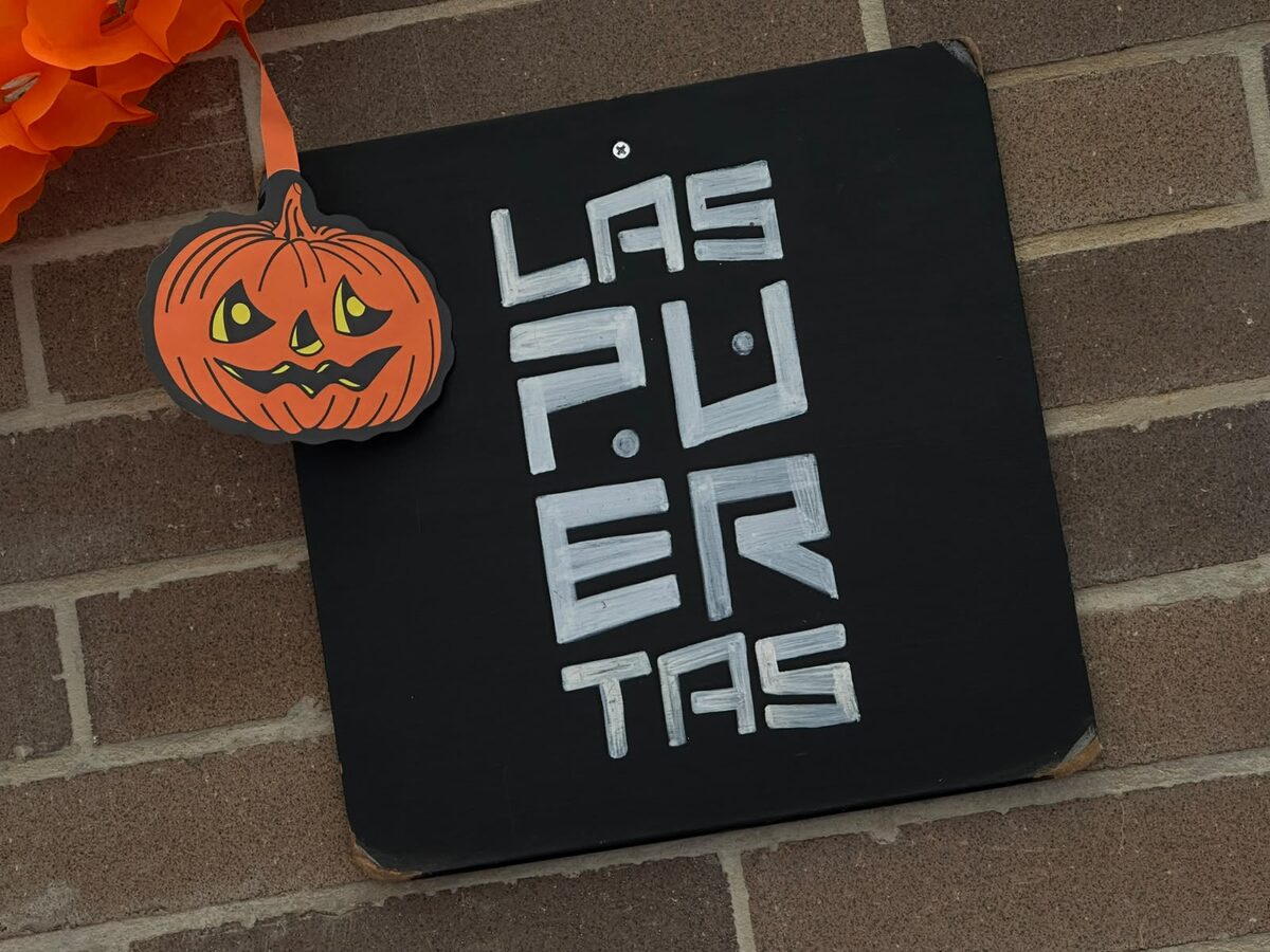 Cartel negro de Las Puertas fijado a una pared de ladrillo a la vista, con una calabaza de papel colgando al lado, decorado para Halloween